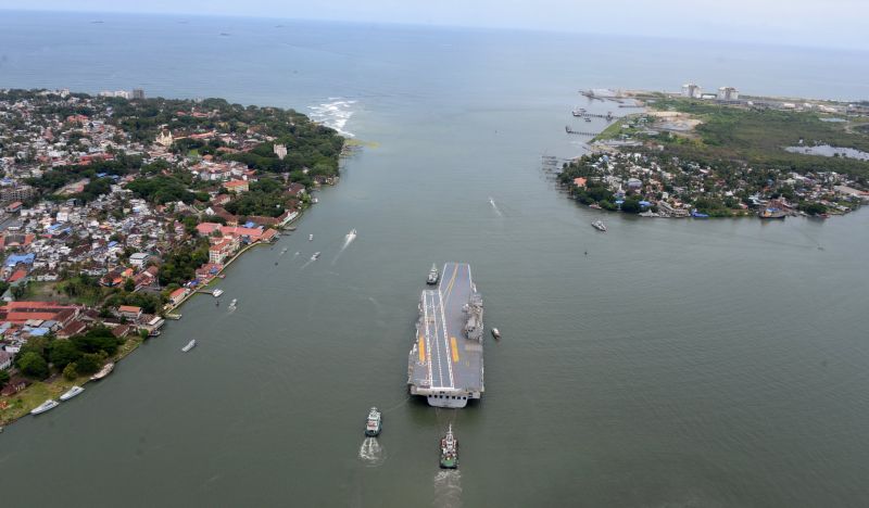 Indegenious aircraft carrier Vikrant sailing through a passage out of the bay with three tiny tugboats following it, as the giant leaves for its sea trials